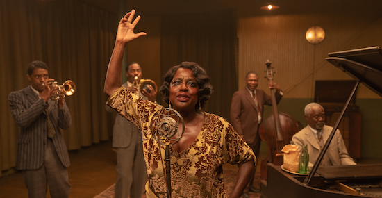 Viola Davis as Ma Rainey stands with her arm upraised performing in front of a microphone at center with other band members in background